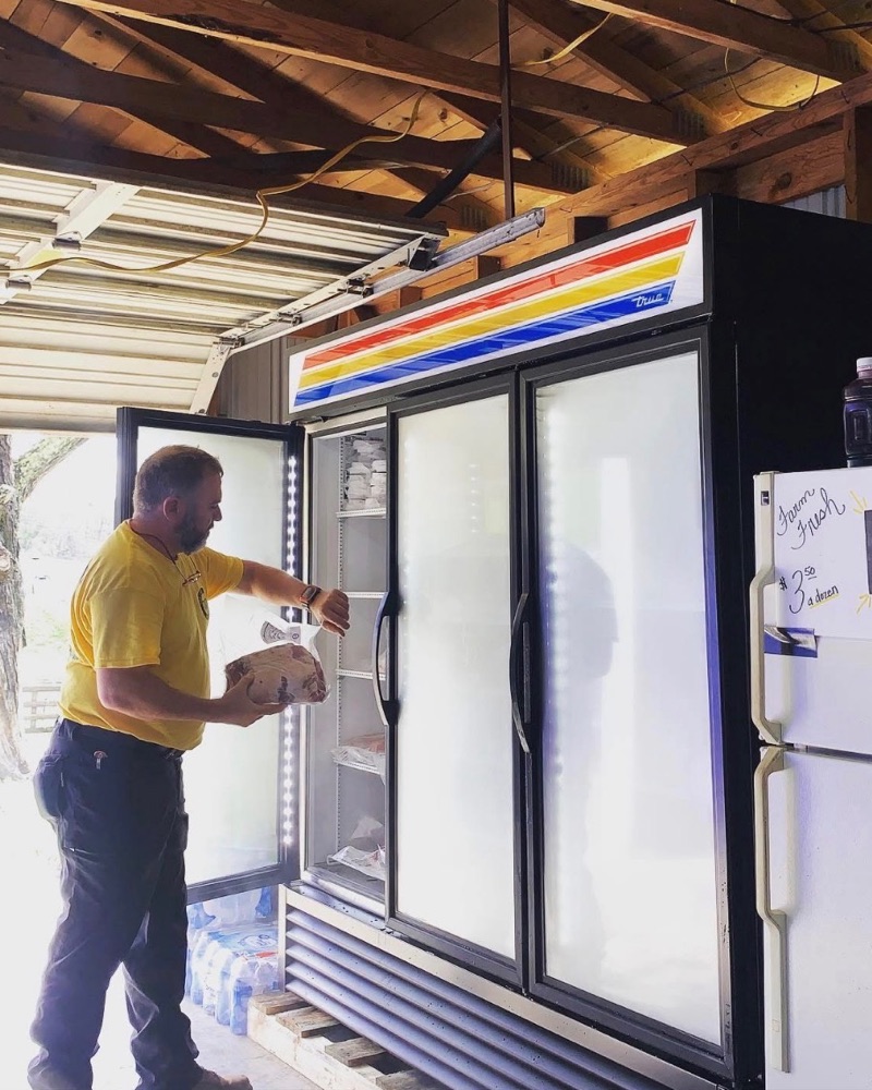 Whitt Shuford loading meat into the single glass-door cooler in the barn where Whitt's Place started, beside a hand-lettered sign for farm fresh eggs