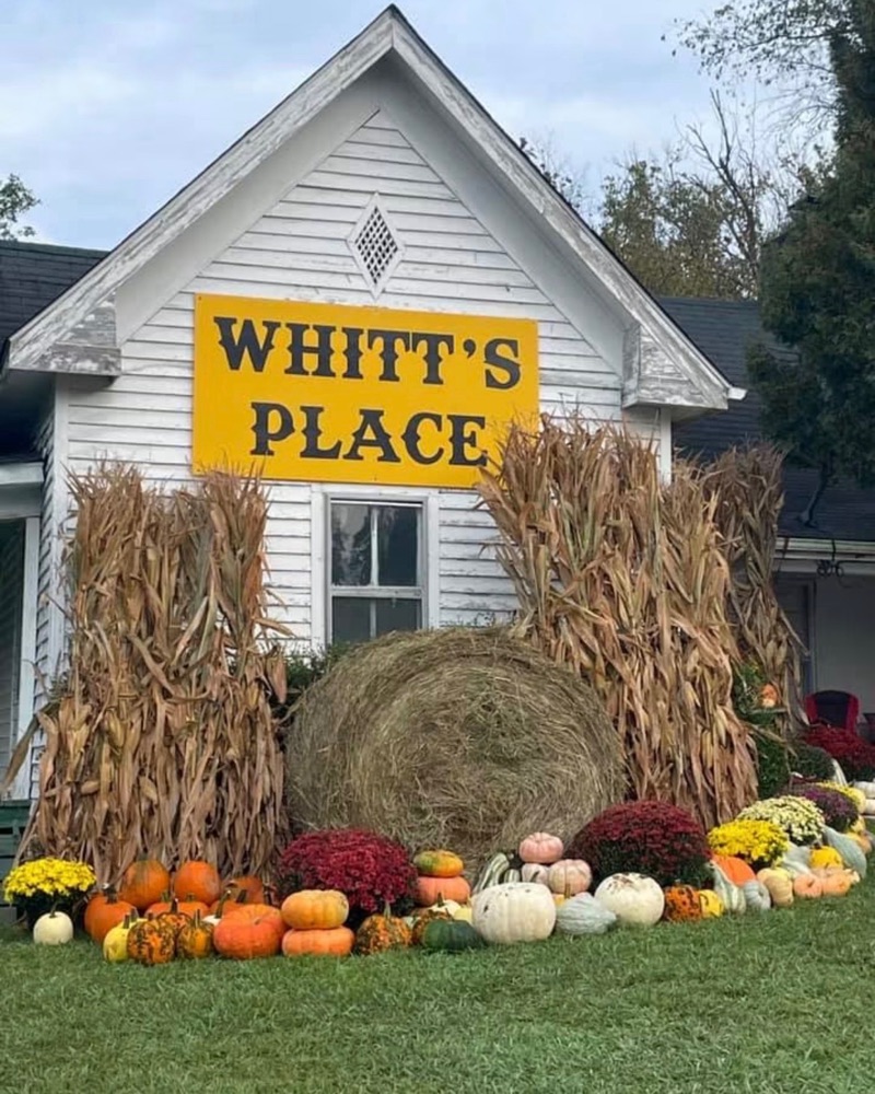 Whitt's Place today, the same farmhouse under a painted sign, with corn shocks, a hay bale, and pumpkins banked across the front